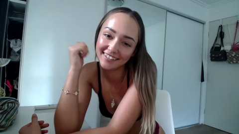 Media: Video of a smiling, light-skinned woman with long, straight brown hair, wearing a black bra, sitting on a white chair in a modern, well-lit room.