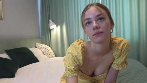 Media: Video of a Caucasian woman with fair skin and blonde hair, wearing a yellow floral dress, sitting on a bed with green curtains in the background.