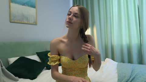 Media: Video of a young Caucasian woman with fair skin and light brown hair in a braid, wearing a yellow off-shoulder dress, sitting on a bed with green pillows and white sheets in a softly lit bedroom.