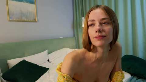 Media: Video of a fair-skinned woman with straight, shoulder-length blonde hair, wearing a yellow off-shoulder dress, sitting on a bed with white sheets and green pillows. The background features a light blue wall and a framed painting.