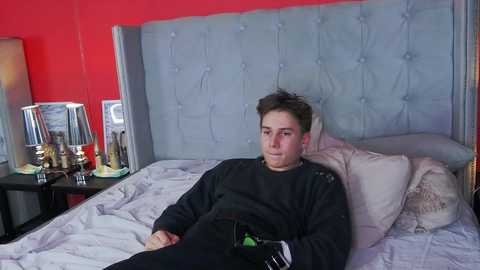 Media: Video of a young man in a black sweatshirt, lying on a neatly made bed with a gray upholstered headboard. The room features a red wall and a bedside table with a lamp and a decorative object.