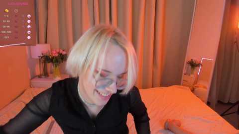 Media: Video of a blonde woman with short hair, wearing glasses and a black blouse, smiling while sitting on a bed with white sheets. The room has beige curtains and a flower arrangement on a nightstand.