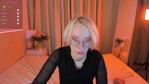 Media: Video of a blonde woman with glasses, wearing a black long-sleeve shirt, sitting on a bed with white sheets, surrounded by orange lighting, against a beige curtain backdrop.