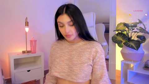 Media: A video of a young woman with long black hair, wearing a cropped knit sweater, standing in a softly lit, modern bedroom with a white chair, pink lamp, and potted plant.