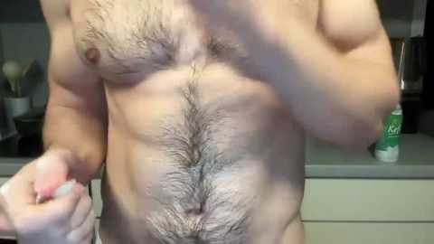 Media: Video of a muscular, light-skinned man with a full chest of dark hair, standing in a kitchen. His right arm is raised, fingers clenched, and he is partially covered by a shadow.