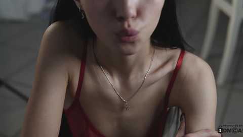 Media: Video of an Asian woman with pale skin and long black hair, wearing a red spaghetti-strap top and silver necklace, blowing a kiss, blurry background.