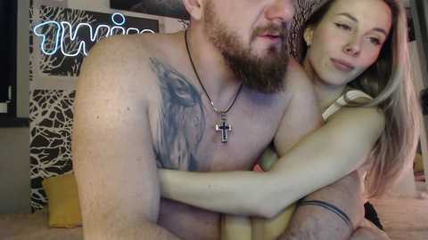 Media: Video of a bearded, muscular man with a tattoo on his chest, embracing a slender, blonde woman with a cross necklace. They are on a bed, with a neon sign in the background.