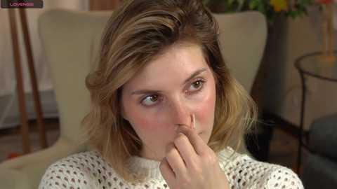 Media: Video of a young Caucasian woman with light brown hair, wearing a white knit sweater, sitting indoors, with a thoughtful expression, finger to her mouth.