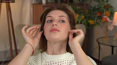 Media: Video of a young woman with fair skin, brown hair, and blue eyes, wearing a white knitted sweater, having her ears adjusted by a hairstylist. Background features a floral arrangement and a green chair.