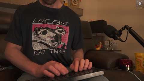 Media: Video of a man in a black \"Live Fast Eat Trash\" T-shirt, typing on a laptop keyboard, against a dimly lit background with a microphone, a yellow container, and a wooden shelf.