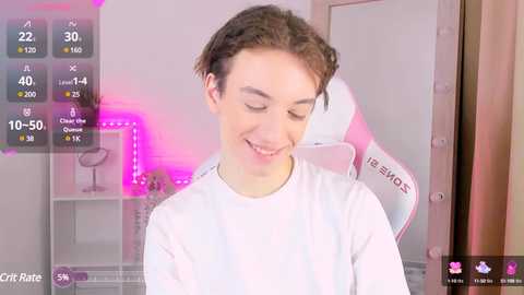 Media: Video of a young, fair-skinned male with short brown hair, wearing a white T-shirt, smiling, in a brightly lit, modern gaming room with a pink and purple LED strip.