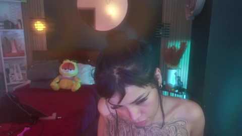 Media: A video of a tattooed woman with dark hair, sitting on a red bed in a dimly lit room. A plush bear and a lamp are visible in the background.