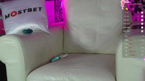 Media: Video of a white leather armchair with a blue vibrator resting on the seat, a white pillow with \"AMSTERBET\" logo, and a pink background, likely taken from a live stream or video chat.