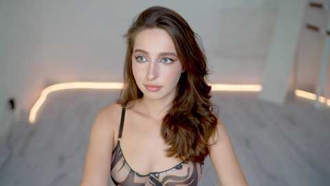 Media: Video of a young woman with long, wavy brown hair, wearing a sheer black lace bra, standing in a modern, softly lit room with white walls and light wooden floors.