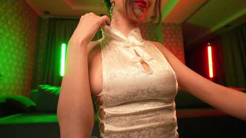 Media: Video of a light-skinned woman in a revealing, shiny white dress with cut-outs, posing in a dimly lit room with green and red neon lights.