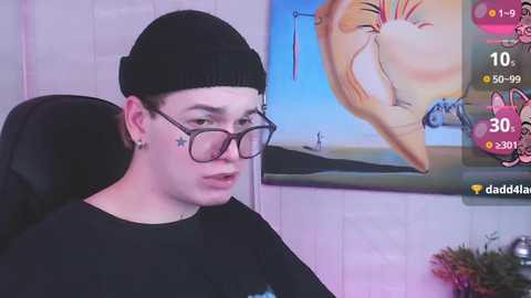 Media: A video of a young, fair-skinned man with glasses and a black beanie, wearing a black shirt, sitting indoors. Background includes a colorful abstract painting and a virtual avatar screen.