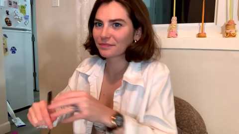 Media: A video of a fair-skinned woman with shoulder-length brown hair, wearing a white button-down shirt, seated indoors. She holds a pen and smiles. Behind her, a fridge with magnets and a window with candles.