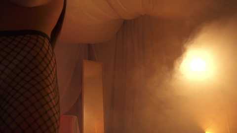 Media: Video of a dimly-lit room with a figure in black fishnet stockings standing in the foreground, casting a shadow. The background features a glowing, warm light source creating a soft, intimate atmosphere.