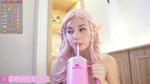 Media: Video of a pale-skinned woman with long, wavy pink hair and pointed elf ears, drinking from a pink \"Starbucks\" straw cup, in a cozy, wooden-paneled room.