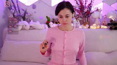 Media: Video of a fair-skinned, dark-haired woman in a pink button-up sweater, holding a glowing, orange object. Background features a white sofa with stuffed animals and a festive, lit-up Christmas tree.