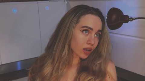 Media: Video of a young Caucasian woman with long, wavy blonde hair, blue eyes, and a surprised expression. She has a slender build and is topless. The background features a white wall and a dark metal light fixture.