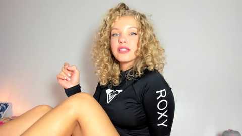 Media: Video of a light-skinned, curly-haired woman with a full face of makeup, wearing a black \"ROXY\" long-sleeve top, sitting on a bed with a white wall backdrop.