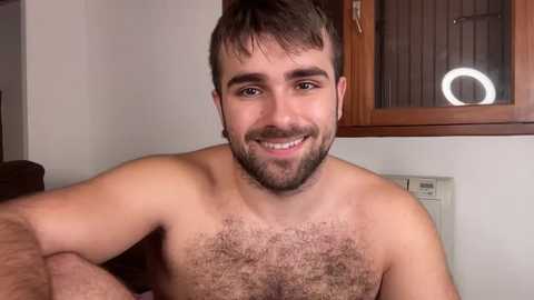 Media: Video of a smiling, shirtless, bearded young man with light skin, short dark hair, and a hairy chest. Background features a wooden-framed window with a circular light and white wall.