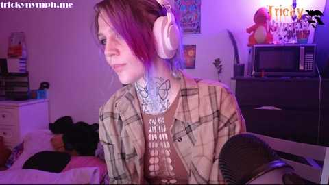 Media: Video of a young woman with fair skin, brown hair, wearing headphones, a plaid shirt, and a lace top, seated in a cluttered room with a purple-lit background.