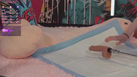 Media: A video of a plush, anthropomorphic rabbit with a pink and white fur, wearing a blue harness, lying on a white bed with pink fur accents. The background features vibrant, abstract art with splashes of teal and red.