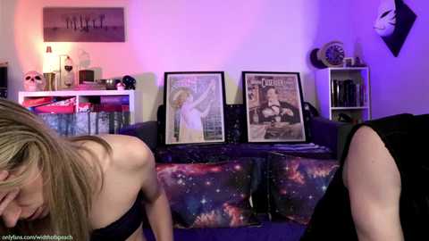 Media: Video of a woman in a black dress, bending over a bed with a space-themed duvet, surrounded by framed magazine covers, in a dimly lit room with purple and pink lighting.