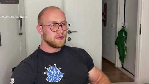 Media: Video of a bald, bearded, light-skinned man in glasses, wearing a black t-shirt with blue basketball logo, standing in a white hallway with green umbrella hanging on wall.