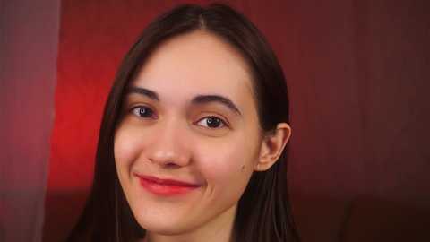 Media: Video of a young woman with long, straight brown hair, fair skin, and a warm smile. She wears minimal makeup, and the background features a soft red gradient.