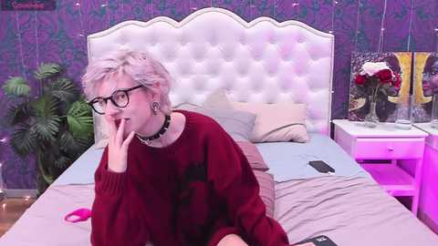 Media: A video of a woman with short, platinum blonde hair, glasses, and a black choker, sitting on a bed with a white tufted headboard, wearing a maroon sweater.