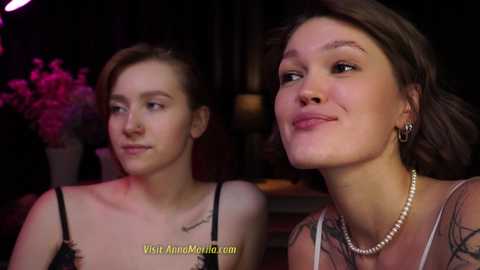 Media: Video of two women: a young Caucasian woman with light skin, dark hair, and a tattoo on her neck, wearing a black top, and a middle-aged woman with medium skin tone, brown hair, and a pearl necklace.