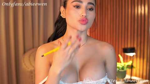 Media: Video of a woman with long dark hair, wearing a strapless white lace dress, applying lipstick with a yellow pencil in a dimly lit room.