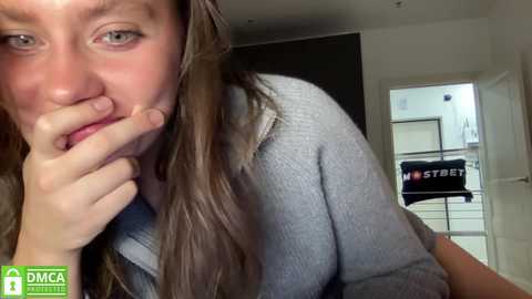 Media: Video of a young woman with long brown hair, wearing a gray sweater, biting her finger, indoors with a \"DCA\" logo in the bottom left corner.