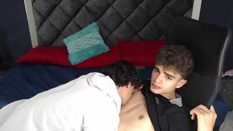 Media: Video of two young men: one shirtless, the other in a black suit, sharing a blanket on a bed with red and turquoise pillows.