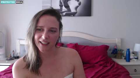 Media: A video of a wet, smiling woman with shoulder-length brown hair, in a pink towel, sitting on a bed with red sheets and white nightstand.