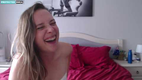 Media: A candid video of a young woman with long, wavy hair, laughing joyfully in bed. She has fair skin and is wearing a white tank top. The room features a white headboard, a pink blanket, and a framed black-and-white photo on the wall.