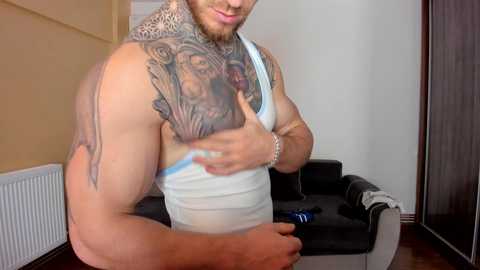 Media: Video of a muscular, tattooed man with a beard, wearing a white tank top, posing in a modern, minimalist room with a dark couch and beige walls.