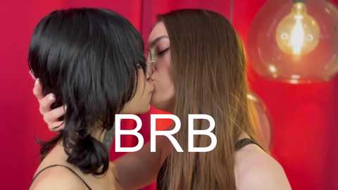 Media: Video of two women kissing passionately, one with long black hair and the other with long brown hair, both in black tank tops. Red background with hanging lightbulb. Text \"BBR\" overlaid.