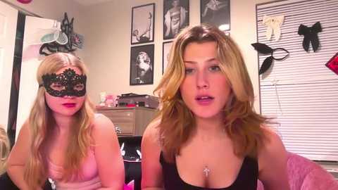 Media: Video of two blonde women in lingerie, one wearing a black lace mask, in a bedroom with framed pictures and clothing on the wall.