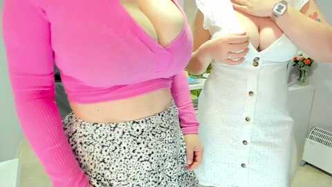 Media: Video of two women: one in a tight, pink crop top and leopard-print skirt, the other in a white lace dress with buttons, standing indoors.