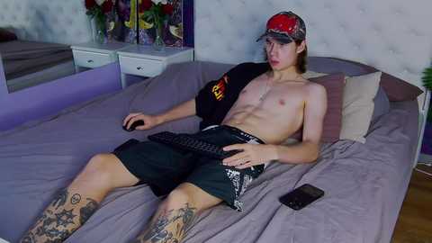 Media: Video of a shirtless, slender young man with tattoos on his legs, wearing a black cap and black shorts, playing a video game on a bed in a purple room.