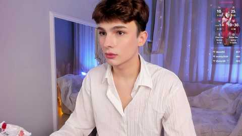 Media: Video of a young, fair-skinned male with short brown hair, wearing a white button-up shirt, standing indoors near a bed with white sheets. Background includes a mirror and a digital scoreboard.