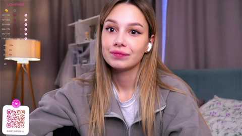 Media: Video of a young woman with long blonde hair, wearing a gray hoodie, white tee, and white earbuds, sitting indoors near a lamp and bed with floral sheets.