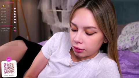 Media: Video of a young Asian woman with long, straight brown hair, wearing a white t-shirt, lying on a bed with purple sheets, looking thoughtful, blurred background.