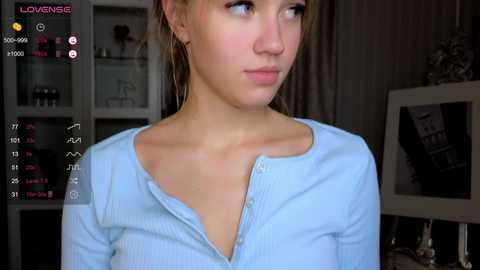 Media: A video of a young woman with fair skin, light brown hair, wearing a light blue, ribbed, button-up shirt, standing indoors. Background includes a white bookshelf and a framed picture.