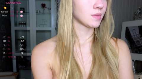 Media: A video of a fair-skinned, topless young woman with long, straight blonde hair, captured in a dimly lit room. The background features a glass cabinet with displayed items, a whiteboard, and a bookshelf.