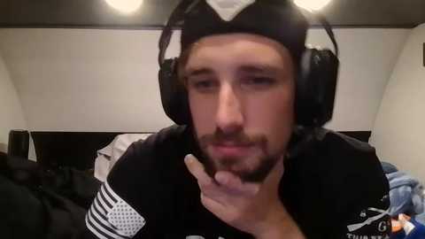 Media: Video of a young man with a trimmed beard and short hair, wearing a black hoodie and oversized headphones, resting his chin on his hand in a casual indoor setting.